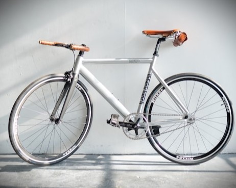 Silver fixed-gear bicycle with brown leather saddle leaning against a white wall in natural light